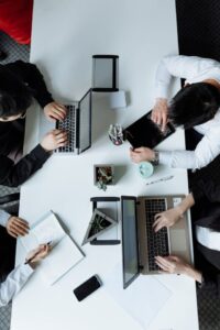 Vista cenital de varias personas trabajando alrededor de una mesa blanca en una oficina, con dos portátiles abiertos, una calculadora, una libreta con notas, un teléfono móvil, bolígrafos y pequeños objetos de escritorio.