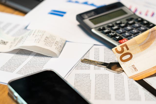 Calculadora negra sobre varios documentos impresos, junto a un billete de 50 euros, una llave metálica, un recibo arrugado y un teléfono móvil en una mesa clara.