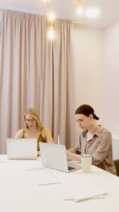 Dos mujeres trabajando con computadoras portátiles en una oficina con cortinas claras y luces cálidas, realizando tareas administrativas vinculadas a la automatización de procesos empresariales