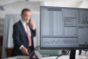 Un monitor de computadora en primer plano muestra un panel de control con gráficos de barras, tablas de datos y métricas financieras como parte de las mejores herramientas de automatización de procesos empresariales. Al fondo, desenfocado, se observa a un ejecutivo con traje y corbata roja hablando por teléfono en una oficina moderna.