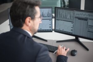 Vista desde la espalda de un hombre de negocios con gafas frente a tres monitores de escritorio que muestran interfaces de software con gráficos de líneas y flujos de información en tiempo real. La imagen ilustra el uso de las mejores herramientas de automatización de procesos empresariales para la supervisión operativa y la gestión de datos en un entorno corporativo.