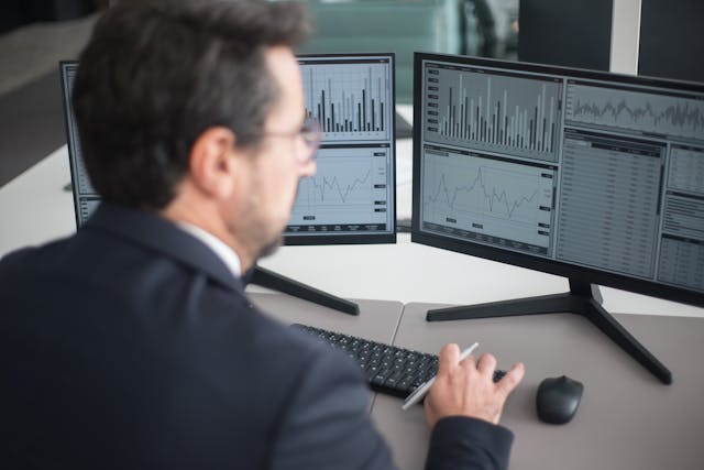 Vista desde la espalda de un hombre de negocios con gafas frente a tres monitores de escritorio que muestran interfaces de software con gráficos de líneas y flujos de información en tiempo real. La imagen ilustra el uso de las mejores herramientas de automatización de procesos empresariales para la supervisión operativa y la gestión de datos en un entorno corporativo.