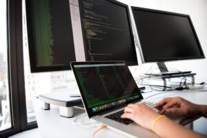 Primer plano de unas manos escribiendo en un portátil que muestra líneas de código en una interfaz oscura. Detrás, dos monitores grandes de escritorio también muestran código de programación complejo. El entorno de trabajo ilustra la implementación técnica de flujos de trabajo con n8n para empresas en una oficina moderna con vistas a la ciudad.