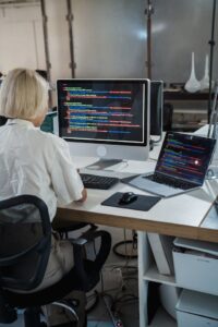 Vista posterior de una mujer con cabello rubio corto sentada en una silla de oficina ergonómica, trabajando frente a dos pantallas que muestran código de programación. La pantalla grande principal está en primer plano, con código visible. Esta configuración de desarrollo de software representa el backend técnico y la programación involucrada al definir que es un sga y cómo se construye este tipo de sistema para la gestión logística.