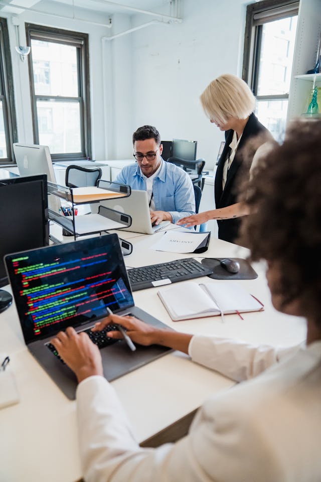 Una vista desde el hombro de una mujer programando en un portátil con código de colores en pantalla, mientras en el fondo, un hombre con gafas trabaja en otro portátil y una mujer rubia de pie le consulta algo. Esta escena de oficina moderna ilustra el trabajo técnico de desarrollo e implementación necesario para entender que es un sga (Sistema de Gestión de Almacenes) en un entorno corporativo.