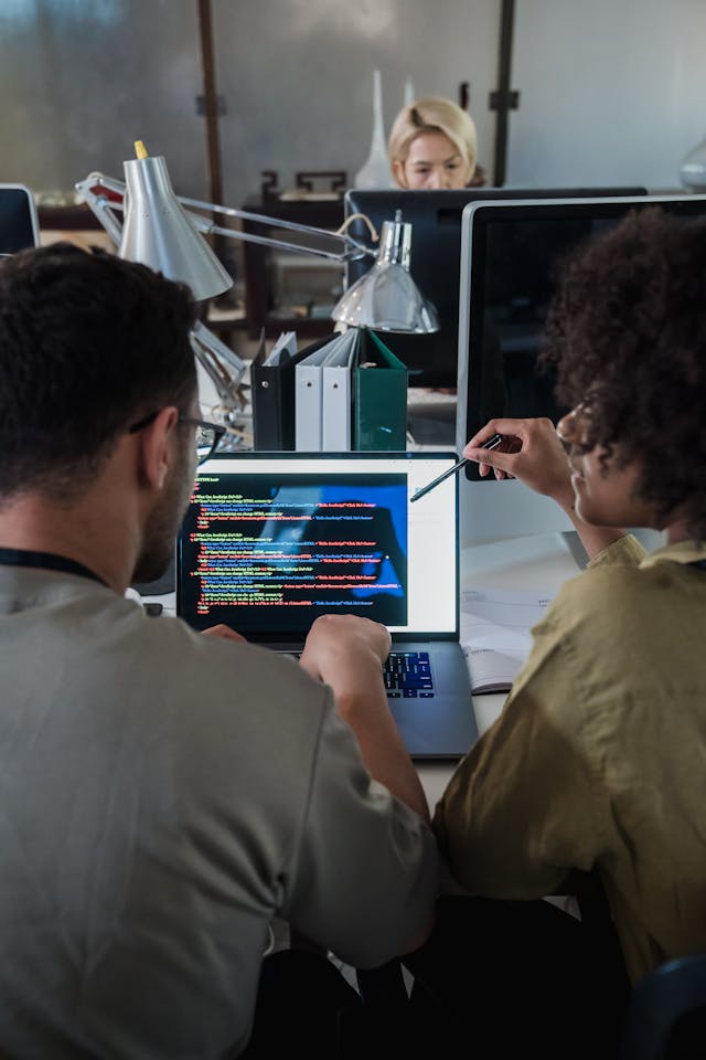 Dos personas están sentadas en un escritorio en una oficina, vistas desde atrás, mirando una pantalla de portátil abierta. Una persona con pelo afro está a la derecha, señalando con un bolígrafo hacia la pantalla, que muestra código de programación con resaltado de sintaxis de colores. La otra persona está a la izquierda, de espaldas. Al fondo, otra persona rubia se ve en otro escritorio. La escena ilustra la colaboración en equipo para definir y revisar un SGA software de gestión de almacén características.