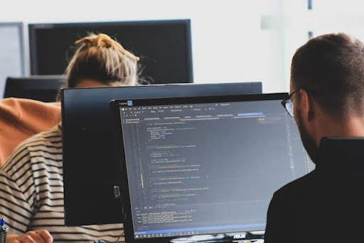 Vista en primer plano desde atrás de un hombre con gafas y camiseta negra mirando un monitor que muestra código de programación en una pantalla oscura. A su izquierda, una mujer con el pelo recogido con un lazo amarillo y una camiseta de rayas horizontales trabaja frente a su propia pantalla en una oficina compartida, un entorno tecnológico ideal para programar las funciones del sga.