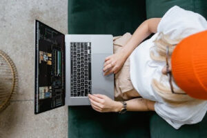 Vista cenital desde arriba de una mujer rubia con un gorro naranja y gafas, sentada en un sofá verde oscuro mientras utiliza un ordenador portátil plateado. Sus manos están sobre el teclado y el trackpad, y la pantalla muestra una interfaz compleja con barras de edición de vídeo y paneles de datos, un escenario de monitorización que ayuda a comprender la diferencia entre erp y sga en la gestión empresarial.