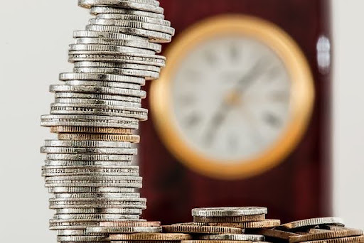 Primer plano de una pila alta y ligeramente inclinada de monedas plateadas y doradas en enfoque nítido, con un reloj analógico de marco de madera desenfocado en el fondo; una representación visual de la relación entre el tiempo y el dinero al analizar como reducir costes en una empresa.