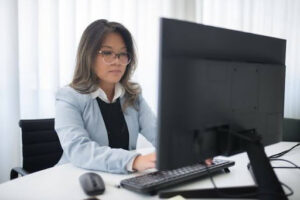 Una profesional con cabello oscuro, anteojos, blusa negra y saco celeste, sentada en una silla de oficina negra. Se encuentra concentrada escribiendo en un teclado negro sobre un escritorio blanco frente a un monitor de computadora, con cortinas blancas iluminadas en el fondo; una representación de las tareas operativas e implementación de herramientas al planificar como digitalizar una empresa.