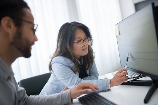Dos profesionales trabajando juntos en un escritorio blanco de oficina. Una mujer de cabello oscuro con anteojos y saco celeste señala la información en la pantalla de un monitor con un bolígrafo, mientras un hombre con barba y lentes la observa atentamente sentado junto al teclado; una escena de trabajo en equipo que ilustra el proceso de como digitalizar una empresa.